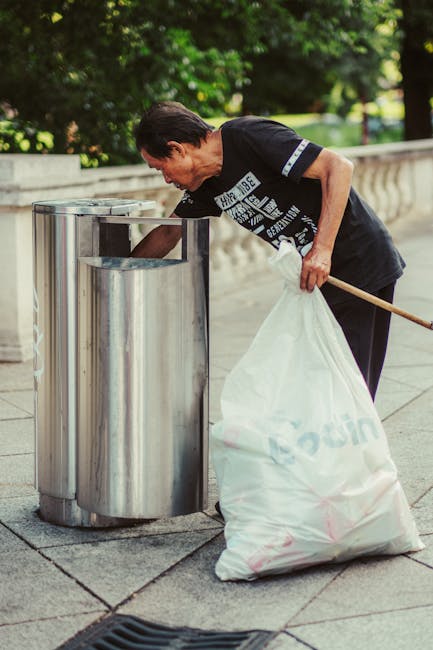 A woman with short dark hair, dressed in a black T-shirt with white printed text, is standing on a paved sidewalk outdoors. She is bending forward, placing a large white plastic bag filled with rubbish into a stainless steel, cylindrical trash bin designed for waste separation. The trash bin has a shiny, reflective surface and an open top, with a second similar bin visible nearby. The surrounding environment includes a stone balustrade on the right, and lush green trees providing shade and a natural backdrop. The scene is lit by natural daylight, creating a clear, neutral atmosphere. This image illustrates an act of independent rubbish disposal, which can be associated with private waste collection services offered by companies like Rubbish Clearance Belgravia, emphasizing proper waste management outside of public council collections. The scene highlights environmentally conscious waste separation and collection in a residential or public park setting, aligning with a professional approach to rubbish removal and the importance of responsible waste disposal.
