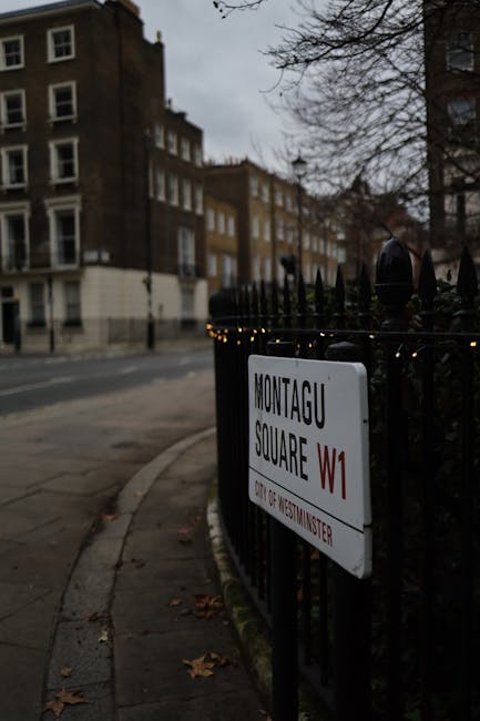 A street scene showing a black wrought iron fence bordering a sidewalk in front of residential buildings with brick facades and multiple windows. The fence has a white street sign attached, reading 'MONTAGU SQUARE W1' with smaller red and black text beneath, indicating a Westminster location. The pavement next to the fence is slightly curved, with a few scattered fallen leaves visible on the ground. In the background, leafless trees and the upper floors of the buildings are under an overcast sky, creating a subdued and cloudy atmosphere. The scene suggests an urban environment with typical Victorian-style architecture, and the fence and sign are indicative of a private or designated area, often associated with property boundaries or small parks within London. Rubbish removal services, such as those offered by Rubbish Clearance Belgravia, often operate in such areas to manage waste from residential properties or local businesses, ensuring proper disposal in line with local regulations and alternative waste handling options within private or public spaces.