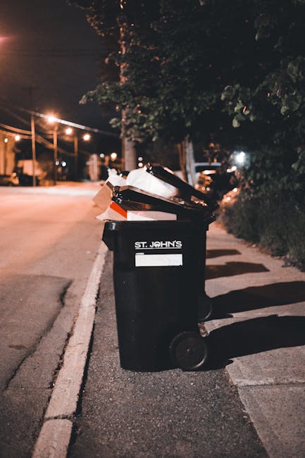 A black wheelie bin labeled with the phrase 'ST. JOHN'S' positioned on a sidewalk at night. The bin is filled with various types of waste, including flattened cardboard boxes, plastic packaging, and miscellaneous rubbish, which are partially visible at the top. The bin is situated adjacent to a curb on a residential street illuminated by warm street lighting, casting shadows on the pavement and nearby greenery. In the background, there are streetlights, a tree, and parked vehicles, indicating an urban setting. The scene reflects an instance of waste disposal typical of private or independent rubbish collection practices, relevant to the context of rubbish removal services provided by companies such as Rubbish Clearance Belgravia. The environment appears quiet, with no people present, emphasizing the bin's role in maintaining waste management outside of official local authority collection, supporting options for private waste handling and on-site clearance.