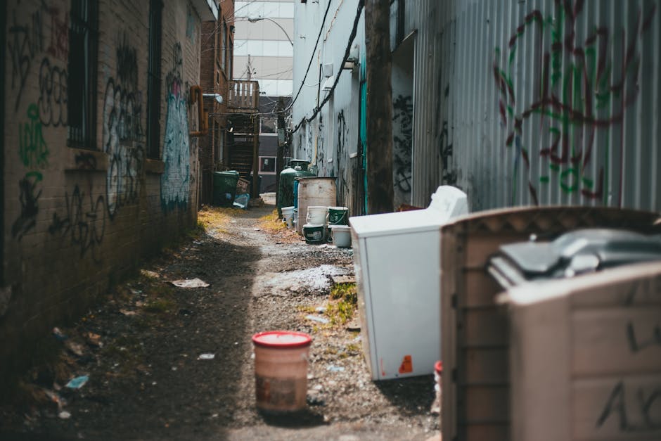A narrow alleyway with a muddy, uneven surface strewn with scattered trash and debris. On the right side, there are various large white and grey household appliances, such as washing machines and refrigerators, some of which are partially obstructing the path. Towards the foreground, a small red and white plastic bucket is visible on the ground. The left side features a brick wall covered with colourful graffiti tags and some small, barred windows. Further down the alley, there are green wheelie bins and other containers, as well as a staircase leading to an upper level at the back. The environment appears weathered and neglected, with patches of moss or mossy growth along the edges of the pathway. Overhead, some wires or cables are loosely hanging, and the natural daylight gives a muted, slightly overcast atmosphere. This scene exemplifies a cluttered urban back alley suitable for private rubbish collection services like those offered by Rubbish Clearance Belgravia, highlighting the need for professional waste removal to tidy such concealed spaces.