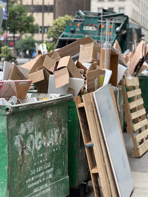 A large green waste bin filled with various cardboard boxes, some flattened and others still in three-dimensional form, along with a few white packaging boxes stacked among them. The boxes are made of brown corrugated cardboard, differing in size and some with printed labels or tape. Leaning against the side of the bin are wooden pallets, some partially visible, with a light natural finish. In the background, there is a street scene with blurred buildings, trees, and a large green waste collection vehicle marked 'Liam' parked nearby. The environment appears to be an urban outdoor setting, with natural daylight illuminating the scene and highlighting the textures of the cardboard and wood. The scene suggests a private rubbish clearance operation, possibly related to an independent waste disposal service, with the debris prepared for collection as part of a rubbish removal process in line with professional waste management practices.