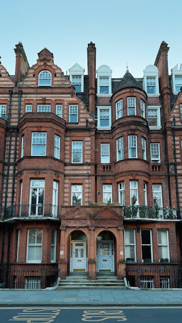 The image depicts a tall, multi-storey Victorian-style residential building constructed from red brick with detailed masonry work around the windows and architectural elements. The building features rounded bay windows on each floor, some with small balconies enclosed by iron railings, and decorative stonework accents highlighting the structure's historic design. The facade includes a central entrance with a pair of white doors, accessed via a short flight of steps and framed by an arched brickwork feature. The top floor has dormer windows set in a mansard roof with slate tiles, alongside brick chimneys rising above. The background shows a clear, pale blue sky with subtle daylight illuminating the building’s textures and colors. The scene is captured from street level, with a paved pavement visible in the foreground. The context suggests an urban setting, possibly involving property management or clearance services like those provided by Rubbish Clearance Belgravia, which handles waste removal from such residential buildings or similar urban structures requiring private rubbish collection or on-site clearance services.
