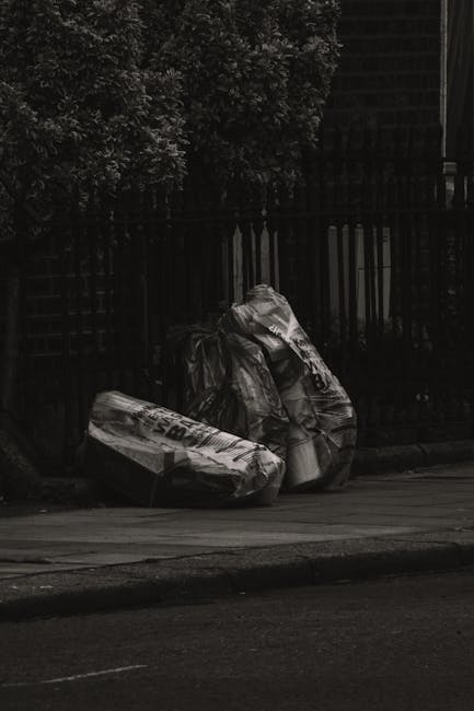 A black-and-white photograph depicts two large bags of waste, approximately filled with rubbish, placed on a paved sidewalk near a dark metal fence with vertical slats. One of the bags appears to be made of plastic, with visible creases and a shiny surface, while the other is more opaque and slightly crumpled. The bags are positioned close to each other, slightly leaning against the fence, with a tree with dense foliage in the background. The scene appears to be in an outdoor urban area, likely associated with private rubbish collection or waste disposal services, as indicated by the presence of these large, possibly commercial-grade rubbish bags on the roadside. The lighting is subdued, possibly reflective of early morning or evening, emphasizing the textures of the waste bags and the surrounding environment. Rubbish Clearance Belgravia might handle such waste, highlighting their role in on-site clearance and alternative waste management methods outside of local authority collection systems.