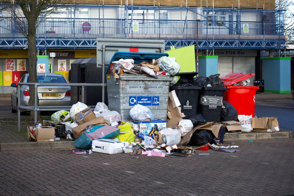 A large accumulation of mixed waste and rubbish bags, including black, white, and red plastic bins, cardboard boxes, paper packaging, and various discarded materials, is piled on a paved sidewalk next to a metal railing in an urban environment. The rubbish appears to be overflowing from a central grey recycling bin labeled for mixed paper and card, with some contents spilling onto the surrounding area. Behind the waste heap, a silver car is parked, and in the background, part of a commercial building with a storefront and signage is visible, along with a tree on the left side. The environment suggests an unmanaged or improperly cleared rubbish collection point, highlighting the importance of alternative waste disposal options such as private rubbish removal services like Rubbish Clearance Belgravia, to prevent rubbish accumulation in communal areas.