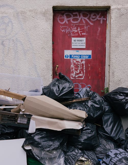A black-and-white photograph depicts two large bags of waste, approximately filled with rubbish, placed on a paved sidewalk near a dark metal fence with vertical slats. One of the bags appears to be made of plastic, with visible creases and a shiny surface, while the other is more opaque and slightly crumpled. The bags are positioned close to each other, slightly leaning against the fence, with a tree with dense foliage in the background. The scene appears to be in an outdoor urban area, likely associated with private rubbish collection or waste disposal services, as indicated by the presence of these large, possibly commercial-grade rubbish bags on the roadside. The lighting is subdued, possibly reflective of early morning or evening, emphasizing the textures of the waste bags and the surrounding environment. Rubbish Clearance Belgravia might handle such waste, highlighting their role in on-site clearance and alternative waste management methods outside of local authority collection systems.